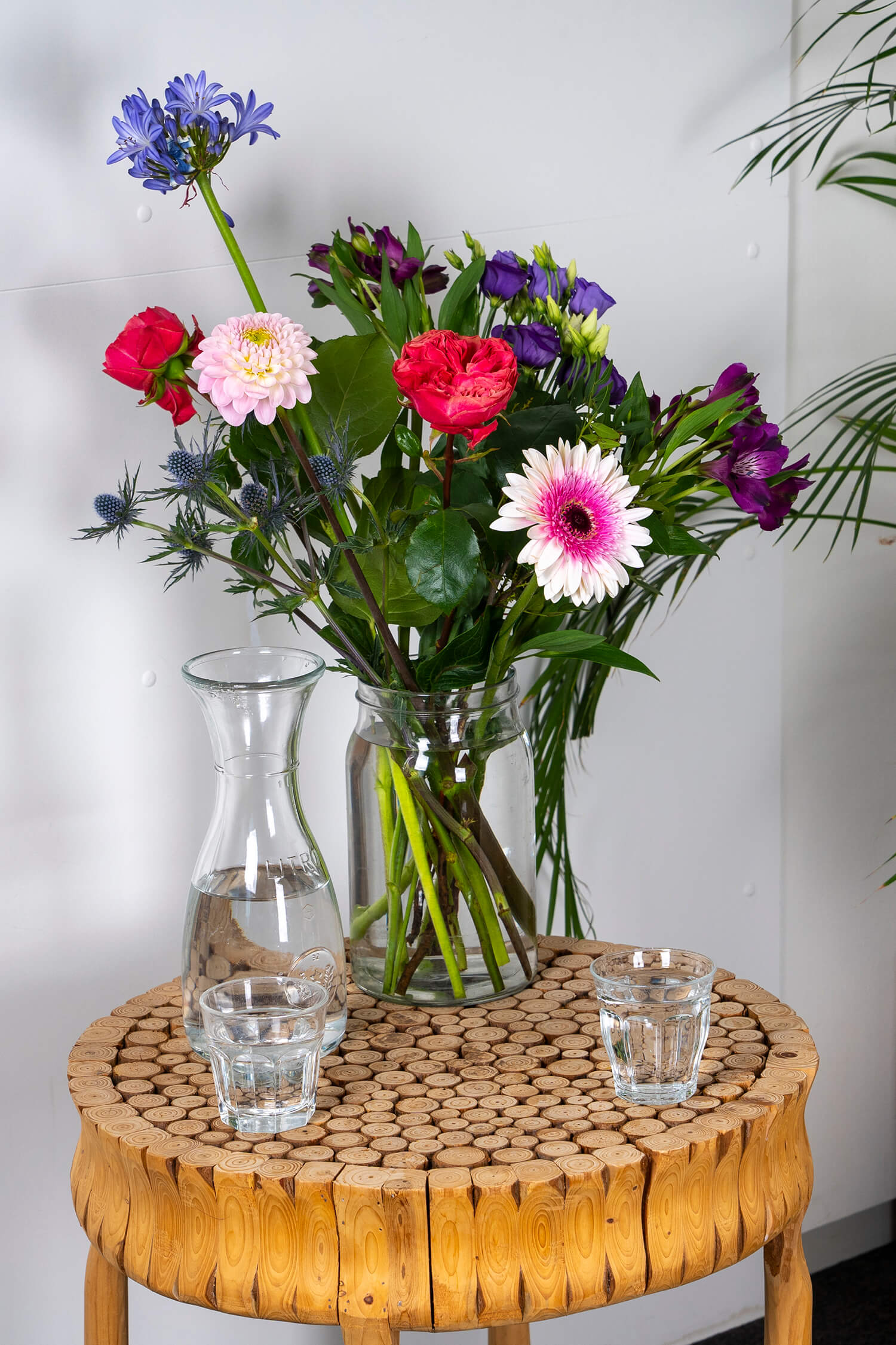 Foto van een bos verse kleurrijke bloemen in een glazen vaas met een karaf water en glazen ernaast.